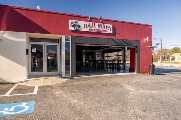 Exterior view of a red and white building labeled "Hail Mary Sports Grille," with large open garage-style doors, patio heaters outside, and a mostly empty parking lot under a clear sky.