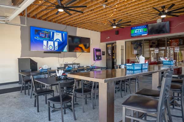 Spacious bar and dining area with high-top tables and chairs, wall-mounted TVs showing sports, exposed wood-beam ceiling with large ceiling fans, and baskets of utensils on the tables.