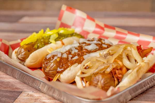 Three hot dogs in buns served in a metal tray with checkered paper, topped with chili and diced onions, pickles and peppers, and sautéed onions with bacon.