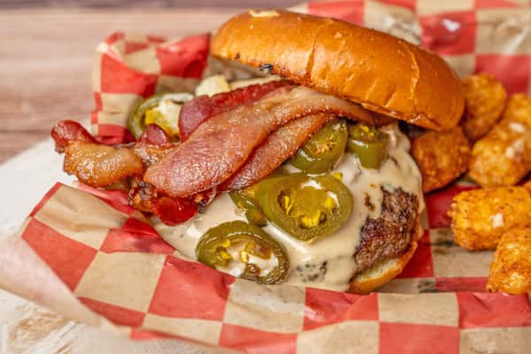Bacon cheeseburger topped with jalapeños and creamy sauce, served with tater tots in a checkered basket.