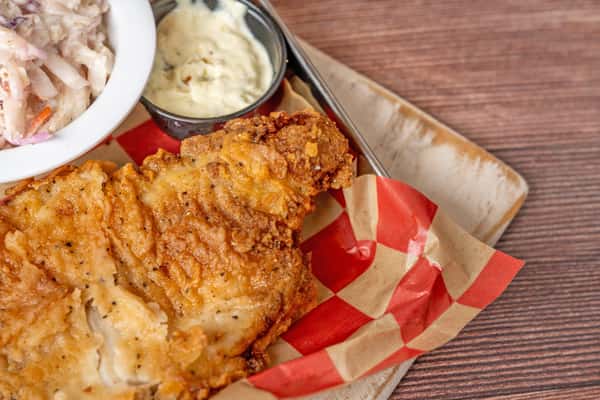 Close-up of crispy fried chicken pieces served on red-and-white checkered paper in a metal tray, with a side of coleslaw and a cup of creamy dipping sauce.