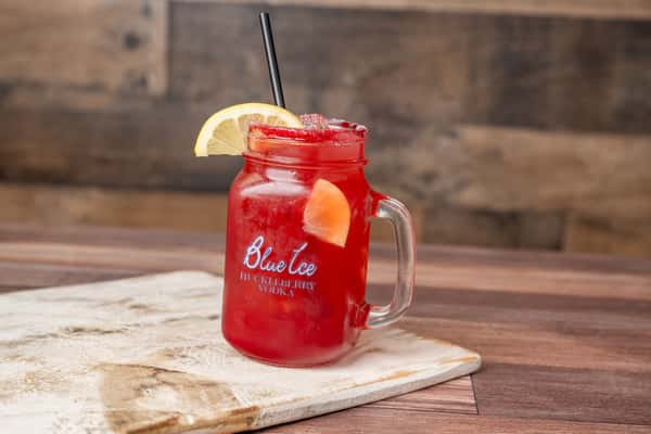 Red cocktail in mason jar with lemon wedge and black straw, Blue Ice Huckleberry Vodka logo on glass, on wood table.