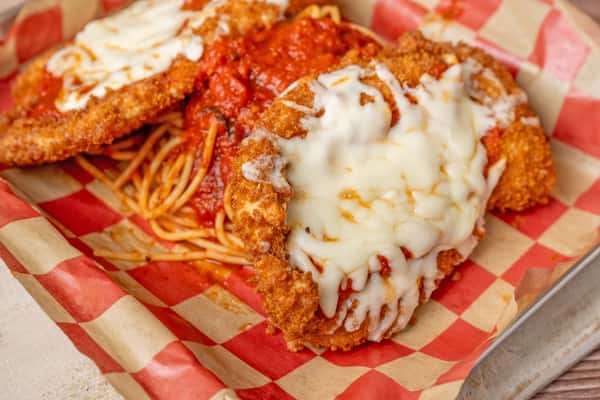 Close-up of a breaded chicken parmesan topped with melted mozzarella and marinara sauce, served over spaghetti in a metal tray lined with red-and-white checkered paper.