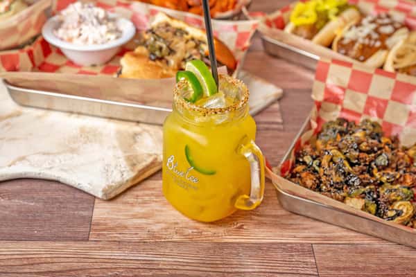 Mango margarita in a mason jar with chili salt rim, lime wedges, ice, and a metal straw on a wooden table.