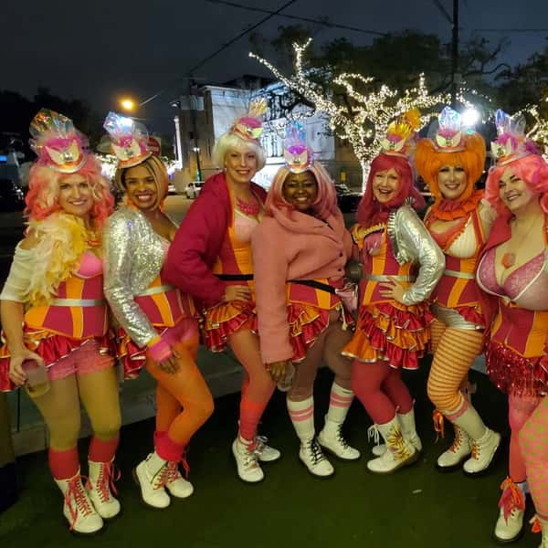 A group of women dressed in vibrant, colorful costumes with pink, orange, and red hues, posing together at night under string lights.