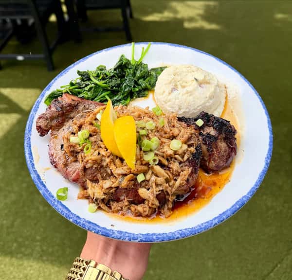 A plate with a grilled pork chop topped with pulled pork, garnished with orange slices and green onions, served with sautéed spinach and mashed potatoes.