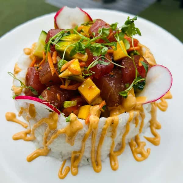 A vibrant poke bowl with fresh tuna, mango, carrots, cucumber, and radish, served on a bed of rice with a drizzle of spicy sauce.