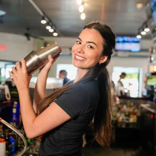 A smiling bartender shaking a cocktail mixer behind the bar at a lively, modern restaurant.