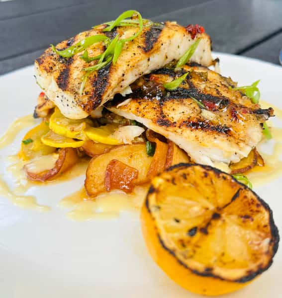 Grilled fish fillets topped with scallions, served over sautéed squash and potatoes with a charred lemon wedge.