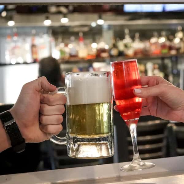 Two hands toasting with drinks at a bar—one holding a frothy mug of beer, the other a red cocktail in a champagne flute.
