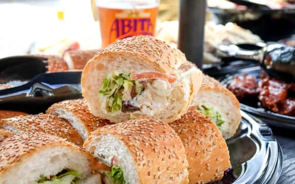 Mini po' boy sandwiches filled with creamy chicken salad, lettuce, and tomato on sesame seed French bread, served on a catering tray.