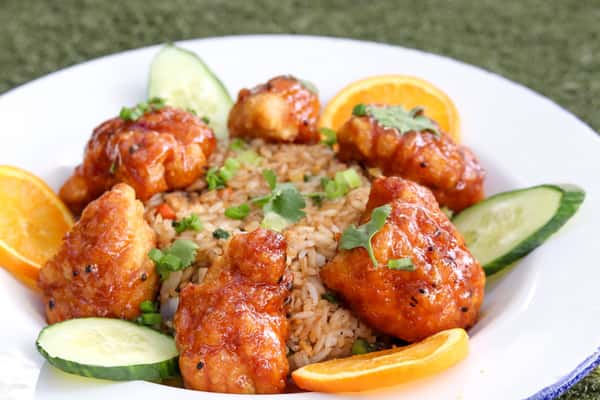 A plate of orange chicken served with seasoned rice, garnished with fresh cilantro, cucumber slices, and orange wedges.