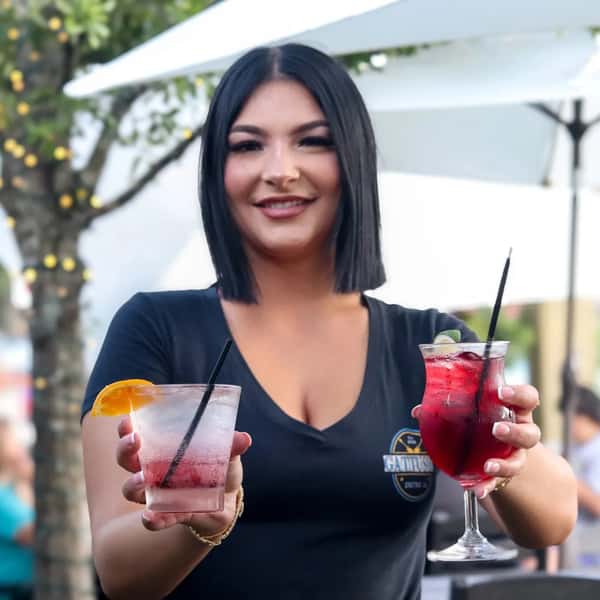 A bartender holding two drinks: one with a slice of orange and the other with a garnish of mint, both served in glasses with straws.