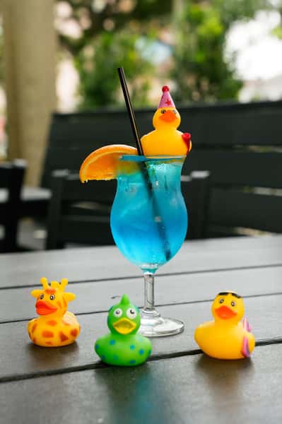 A vibrant blue cocktail garnished with an orange slice and a rubber duck, surrounded by colorful rubber duck toys on a patio table.