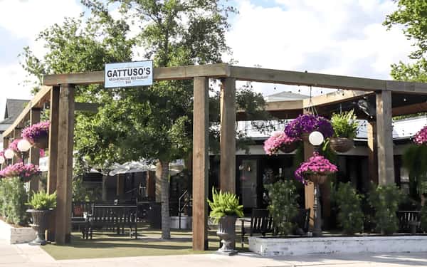 The entrance to Gattuso's Neighborhood Restaurant and Bar, featuring a sign above a wooden structure, with outdoor seating and greenery in the background.