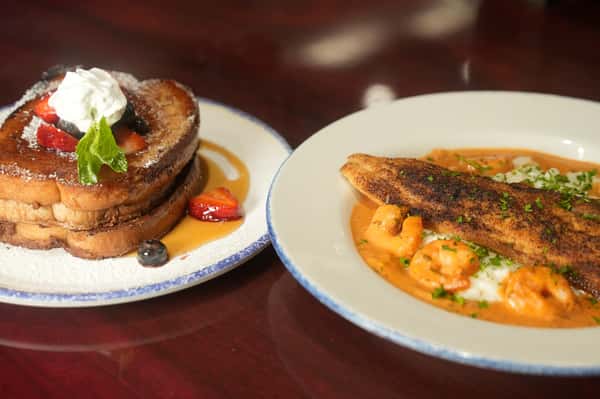 A plate of French toast topped with berries, whipped cream, and syrup, alongside a plate of blackened fish with shrimp in a creamy sauce over rice.