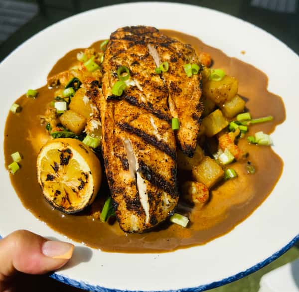A grilled fish fillet served with sautéed vegetables, a grilled lemon, and a rich brown gravy, topped with green onions.