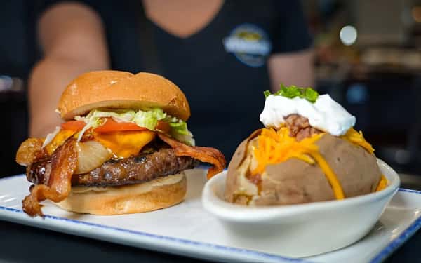 A bacon cheeseburger with lettuce, tomato, cheddar cheese, and grilled onions, served with a loaded baked potato topped with sour cream, shredded cheese, and green onions.
