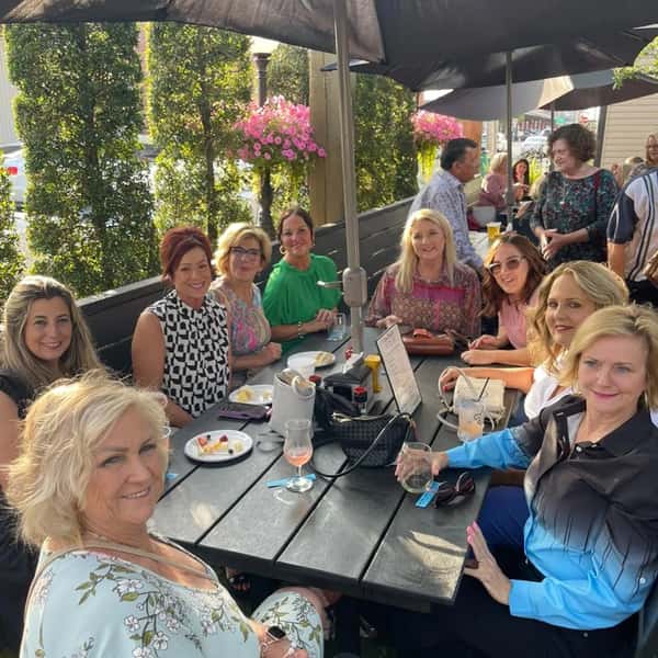 A group of women enjoying food and drinks at a long outdoor table under umbrellas, surrounded by greenery and flowers.