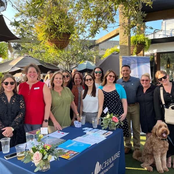 A group of people posing for a photo at an outdoor event, standing in front of a table with a floral arrangement and informational materials. There's a dog at the front, and the background shows umbrellas and greenery.