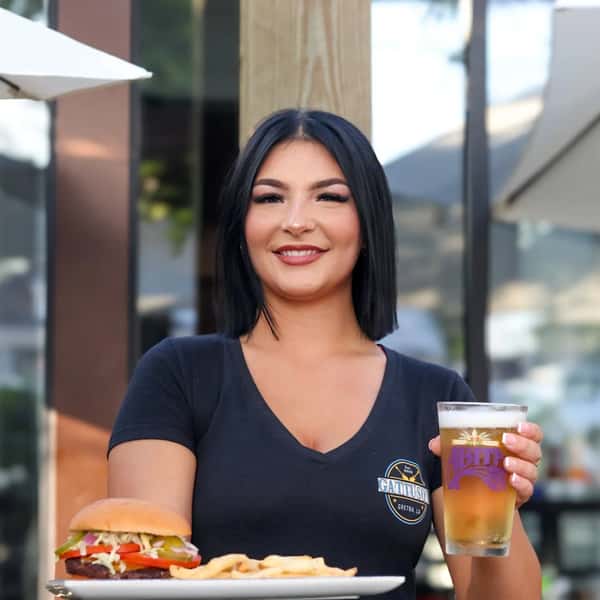Smiling server holding a glass of Abita beer with a plate of fries and a burger at a restaurant patio.