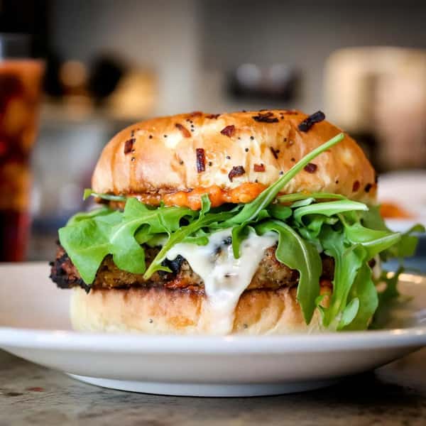 A delicious sandwich featuring a grilled patty topped with arugula and melted cheese, all nestled in a toasted onion bun with a tangy sauce.