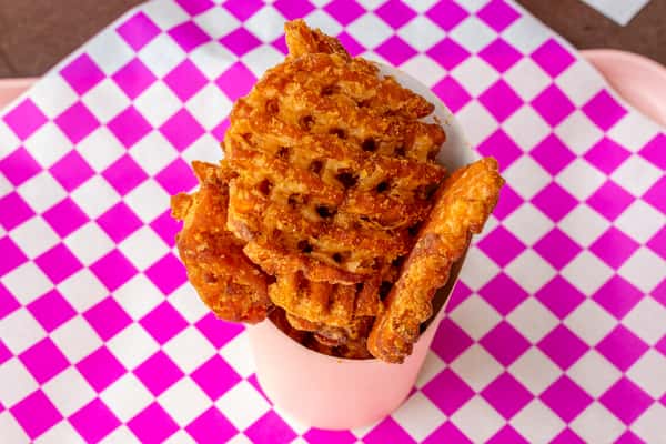 Crispy golden-brown waffle fries in a light pink paper cone on a vibrant pink and white checkered background.