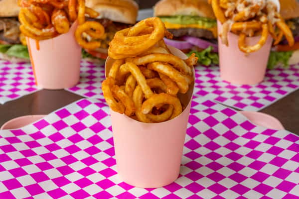 Crispy golden curly fries in light pink cups served on vibrant pink and white checkered paper with burgers in the background.