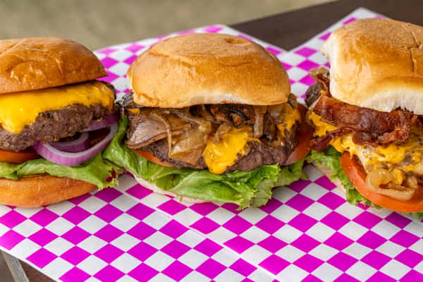 Three delicious cheeseburgers with various toppings like bacon and sautéed onions, served on pink and white checkered paper.
