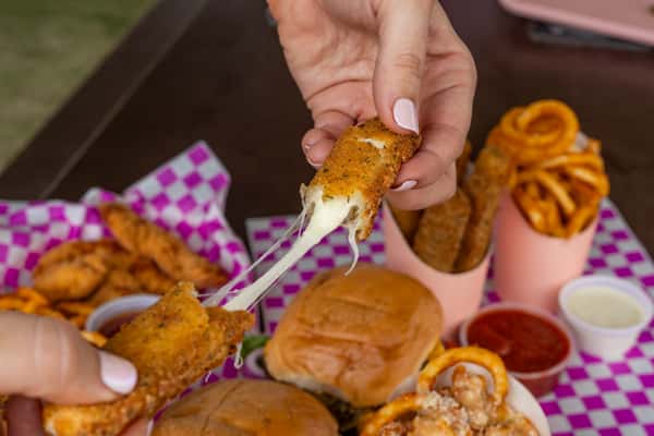 Hands pulling apart a golden mozzarella stick with a long cheese stretch over burgers and curly fries on pink checkered paper.