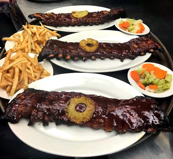 three full racks of ribs topped with barbecue sauce and a pineapple slice. French fries and mixed vegetables on the side.