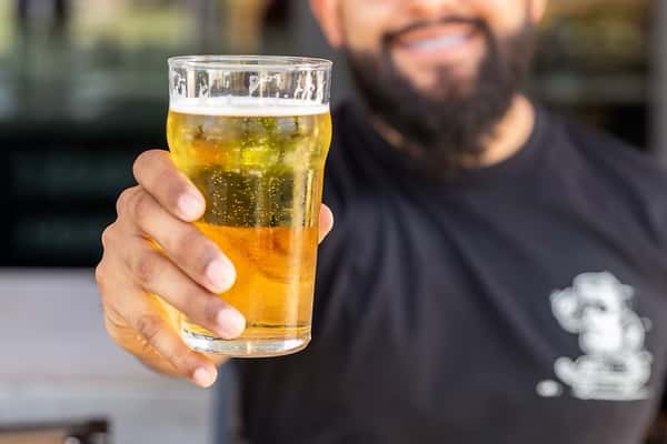 Man holding a pint of golden beer with bubbles, smiling in the background while wearing a black shirt with a white logo.