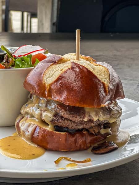 A pretzel bun burger with melted cheese sauce and beef patty served with a side salad and pickle spear on a white plate.