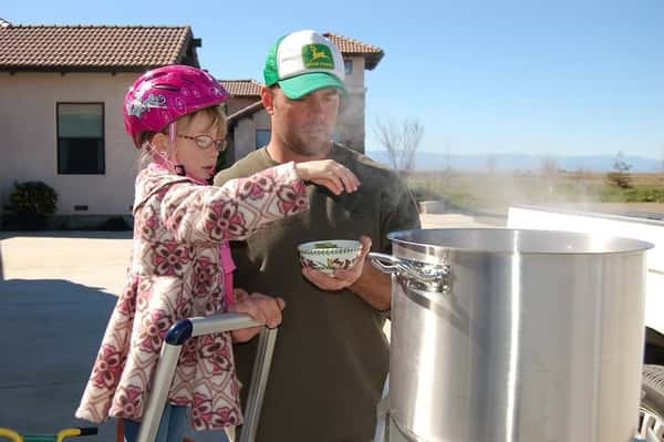 Man holding a young girl adding ingredients into a steaming outdoor brew kettle on a sunny day.