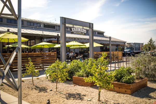 Farmers Brewing Co. exterior with patio seating, green umbrellas, string lights, and raised garden beds in the foreground.