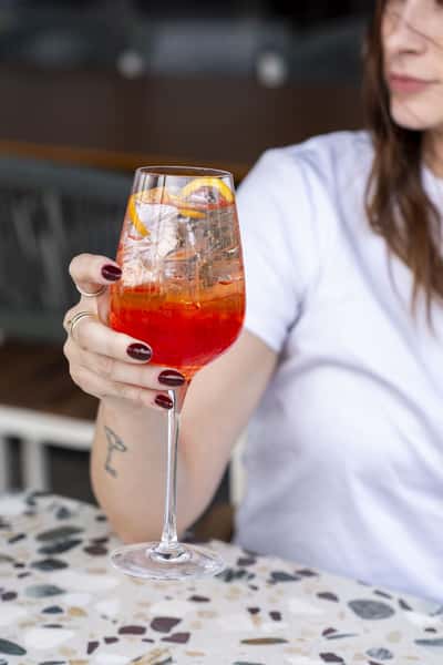 A hand holds a vibrant orange-red cocktail garnished with a dried citrus slice, served in a stemmed glass with a bright and refreshing presentation.