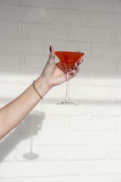 A hand holds up a vibrant red cocktail in a martini glass, showcasing its bold color against a clean white tiled background.