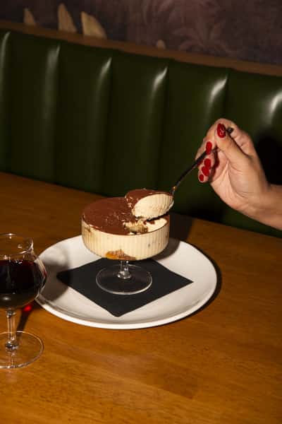 A decadent dessert served in a coupe glass, topped with a crisp pastry and dusted with powdered sugar. A hand sprinkles finishing touches over the dish beside a glass of red wine.