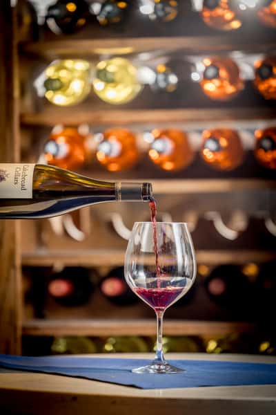 Red wine being poured from a Cakebread Cellars bottle into a glass, with wine bottles displayed on wooden shelves in the background.