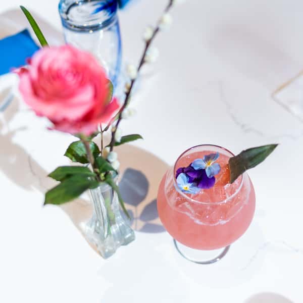 Pink cocktail with ice, edible flowers, and a leaf garnish on a white table next to a vase with a pink rose and willow.