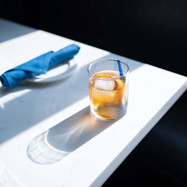 Glass of iced cocktail with a stirrer on a sunlit marble table, next to a plate with a neatly folded blue napkin.