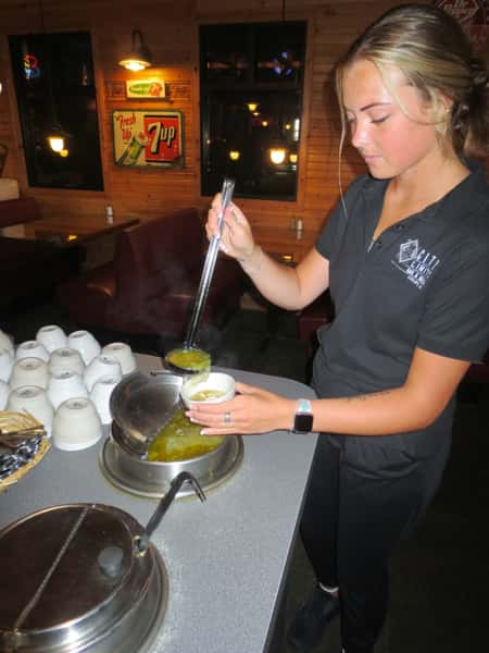 Waitress in black uniform ladles steaming soup at City Limits Bar & Grill, with white bowls, rustic wood paneling and vintage 7-Up sign in the background.