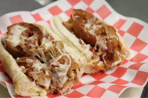 Toasted hoagie sandwich filled with melted cheese, crispy onion rings, diced bacon, and grilled chicken on checkered paper.