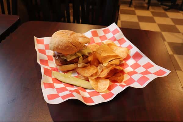 Crispy chicken sandwich with cheese and veggies, served with potato chips and a pickle on red-and-white checkered paper.