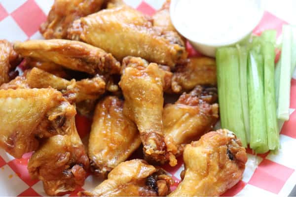 Golden fried chicken wings served with celery sticks and a side of ranch dressing on red and white checkered paper.