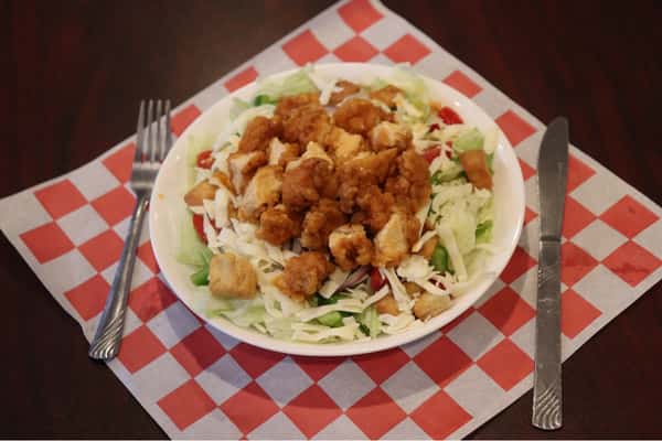 Crispy fried chicken salad served on a bed of shredded lettuce, tomatoes, green peppers, shredded cheese, and crunchy croutons. Presented in a bowl with a red-and-white checkered paper underneath, accompanied by a fork and knife.