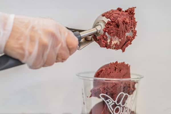 A gloved hand is scooping a thick, dark reddish-purple açaí blend into a clear branded "Alohana" cup, preparing the base for an açaí bowl.