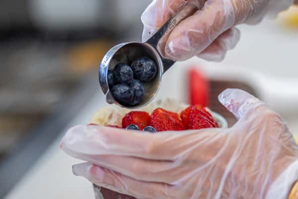 This image shows a gloved hand using a 1/8 measuring cup to add fresh blueberries to the top of an acai bowl already topped with strawberries and banana slices.