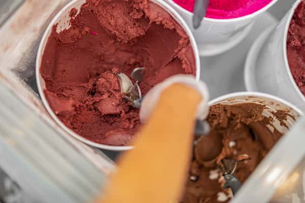 A gloved hand scoops dark red acai sorbet from a container in a freezer, surrounded by other vibrant sorbet flavors like pitaya (pink) and chocolate.