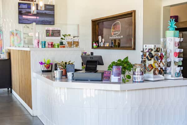 ALOHAHA counter with a register, colorful cups, keychains, plants, and a "Serving San Elijo Since 2020" sign in the background.